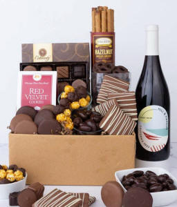 A gift box overflowing with treats including an assortment of chocolate-covered items like oreoes, almonds, graham crackers and pretzels along with red velvet cookies, hazelnut wafers, an 8 piece box of chocolates and a bottle of red wine.